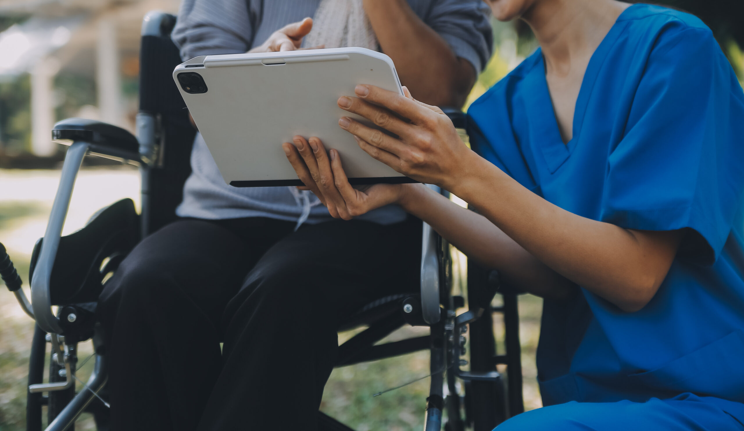 Eine junge Pflegekraft kniet neben einen älteren Frau im Rollstuhl und hält ihr ein Tablet hin.